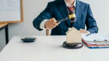 A lawyer for home purchase in a suit holds a gavel above a model house on a desk with a calculator, clipboard, and paperwork—symbolizing the legal steps in buying your home.